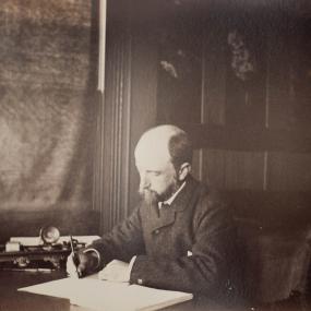 Henry Adams, seated at a desk in a dark panelled room, writing, with large books stacked behind him. Photograph by Marian Hooper Adams, 1883 . Massachusetts Historical Society.