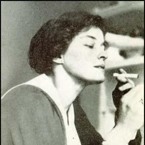 Black and white photograph of Enid Bagnold in profile from the shoulders up. In one upraised hand she holds a cigarette. She wears a dark-coloured shirt with white around the collar and cuffs.