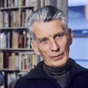 Colour photo of Samuel Beckett in 1977, looking straight at the camera. He has grey hair and blue eyes, and standsin front of bookshelves, wearing a polo-necked shirt and heavy navy sweater.