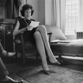 Black-and-white-photo of Patricia Beer sitting in a chair reading her poetry at a festival held at the Royal Court Theatre, London, July
            1963.