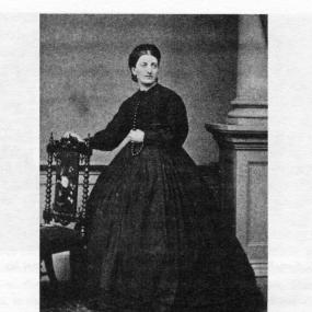 Black and white photograph of Isabella Beeton, standing with her head turned towards the camera, and one hand resting on an ornate chair. She is wearing a black dress with a voluminous skirt and a long, dark beaded necklace.
