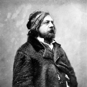 Photograph of Théophile Gautier by Nadar, c. 1856. Gautier stands in profile, three-quarter length, looking to the right, wearing an astrakhan coat and apparently an ethnic hat.