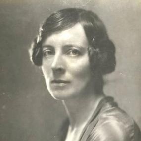 Black and white, head-and-shoulders photograph of Mary Agnes Hamilton, taken from the side but turning her face to the camera. Her blouse or jacket looks silky; her dark hair is in coils over her ears.