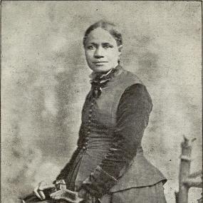 Black and white three-quarter-length photo of Frances E. W. Harper, frontispiece to an edition of her "Poems", Philadelphia, 1898. She is standing, her hair parted in the middle and pulled back; she looks straight forward, direct but dreamy. She is wearing a long skirt and sleeveless top with a shirt under it, with bows at the collar. Her hands rest on a chairback.