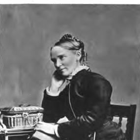 Low resolution black and white three-quarter-length photo of Frances Ridley Havergal sitting on a chair, Her hair is braided in a crown on top of her head. She is wearing a dark, long-skirted dress and jacket with white at the throat and wrists. She sits at a table with two drawers, with a basket like a jewel box on the table and a fat book in her lap. Underneath the image her signature is reproduced: "Frances R. Havergal".