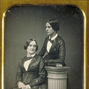 Black and white photograph of Matilda Hays (standing on the right) with Charlotte Cushman (sitting on the left). Matilda Hays is wearing a fancy vest and skirt, with a bowtie around her neck. Her hair is parted in the middle and tied back. Her left arm is resting on a book on top of a doric pillar with floral design on the sides near the top. Charlotte Cushman is wearing a very similar outfit except her skirt has some striped design on it. Her hair is parted in the middle, but it is tied up in a double bun.