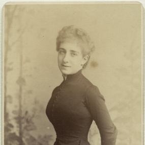 Black and white photograph of Ada Leverson, standing with her hands behind her back. She is wearing a dark dress with a high collar, long sleeves, a broad skirt, and a row of buttons up the front. Her hair is short and curly. Below her photograph are the words: "Elliot and Fry, 55 and 56, Baker St London. W", typed in all capitals.