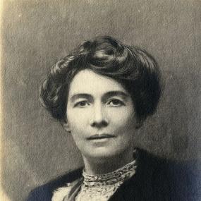 Photograph of a painting of Emmeline Pethick-Lawrence. She is depicted from the shoulders up, wearing a dark jacket over a pale, choker-necked shirt. Her dark, luxuriant hair is pinned on top of her head. Her expression is serene.