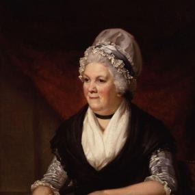Painting of Sarah Trimmer by Henry Howard, exhibited 1798. She sits at a table bearing books (including a bible), inkwell and spare quill pens, holding one pen and some sheets of paper (as well as her spectacles). Her grey hair is mostly covered by a white mob cap with grey ribbon round it; she wears a checked dress and a black shawl with white neckerchief and black neckband. She is smiling slightly. National Portrait Gallery.