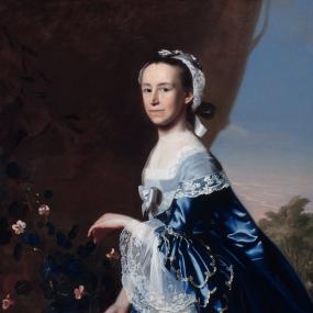 Painting of Mercy Otis Warren by John Singleton Copley. She stands in an outdoor background of trees and flowers, holding one hand out, and wearing a deep blue gown with quantities of lace at the wrists, frills and more lace elsewhere, and a silver bow in the centre of the bodice. She has a lacy white cap tied with a silver ribbon on her brown hair.
