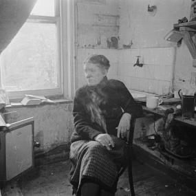 Black-and-white photo of Anna Wickham in her cluttered kitchen, originally published to accompany "Anna Wickham: A Poetess Landlady" in
            Picture Post, 1946. Looking off-camera, she is seated on a chair and smoking a cigarette. 