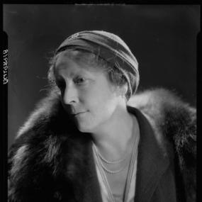 Black and white, head and shoulders photograph of E. H. Young by Howard Coster, 1932. She is in semi-profile, wearing a skullcap-like hat, fur-collared coat, gloves, and a double string of pearls. National Portrait Gallery.